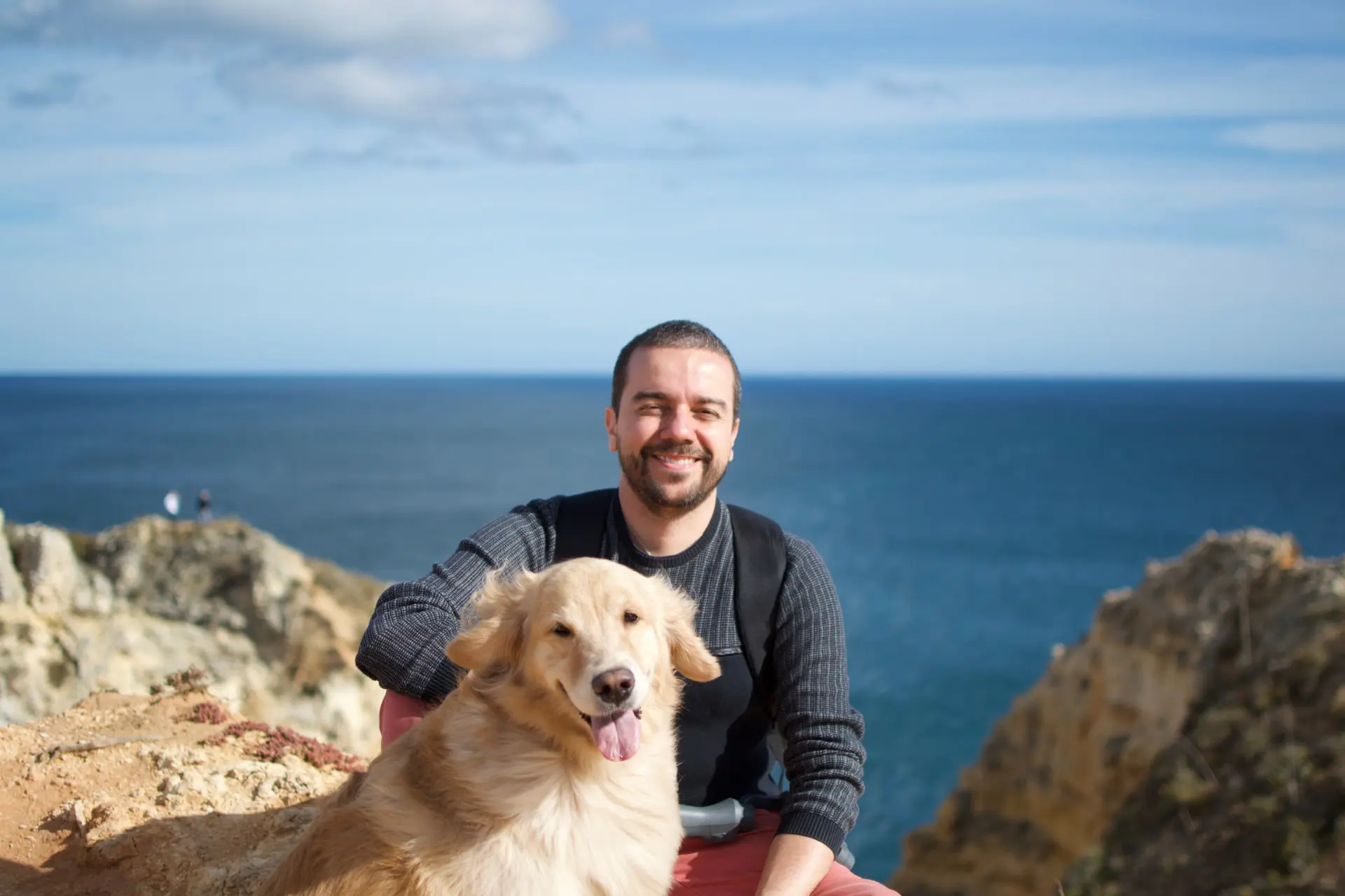 Leandro Sosi sorrindo abraçado à Julieta, sua Golden Retriever, em um mirante à beira-mar